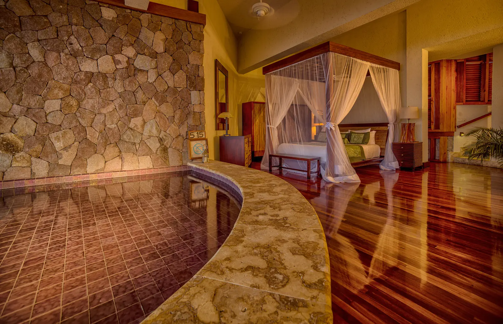 A Star sanctuary: the four-poster bed with its net, a polished timber floor, a stone wall and the pool reaching into the room.