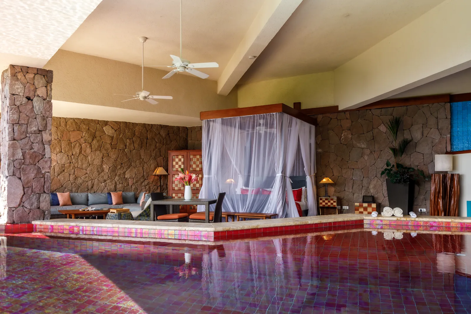 A Sun sanctuary: the four-poster bed under its white net, a sitting area, ceiling fans and the red-tiled pool filling the foreground.