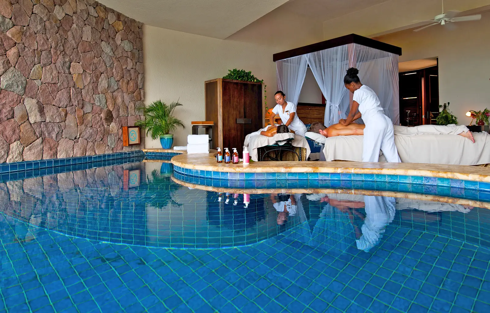 A couple’s massage inside a sanctuary: 2 therapists in white, the tables beside the four-poster bed and the blue-tiled pool in the foreground.