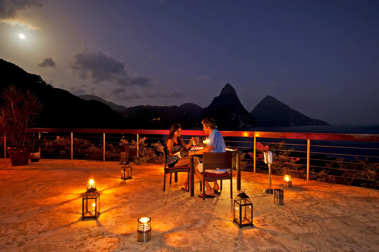 Dinner for 2 on the terrace at night: one table, lanterns on the floor, the moon among clouds and the outline of the Pitons.