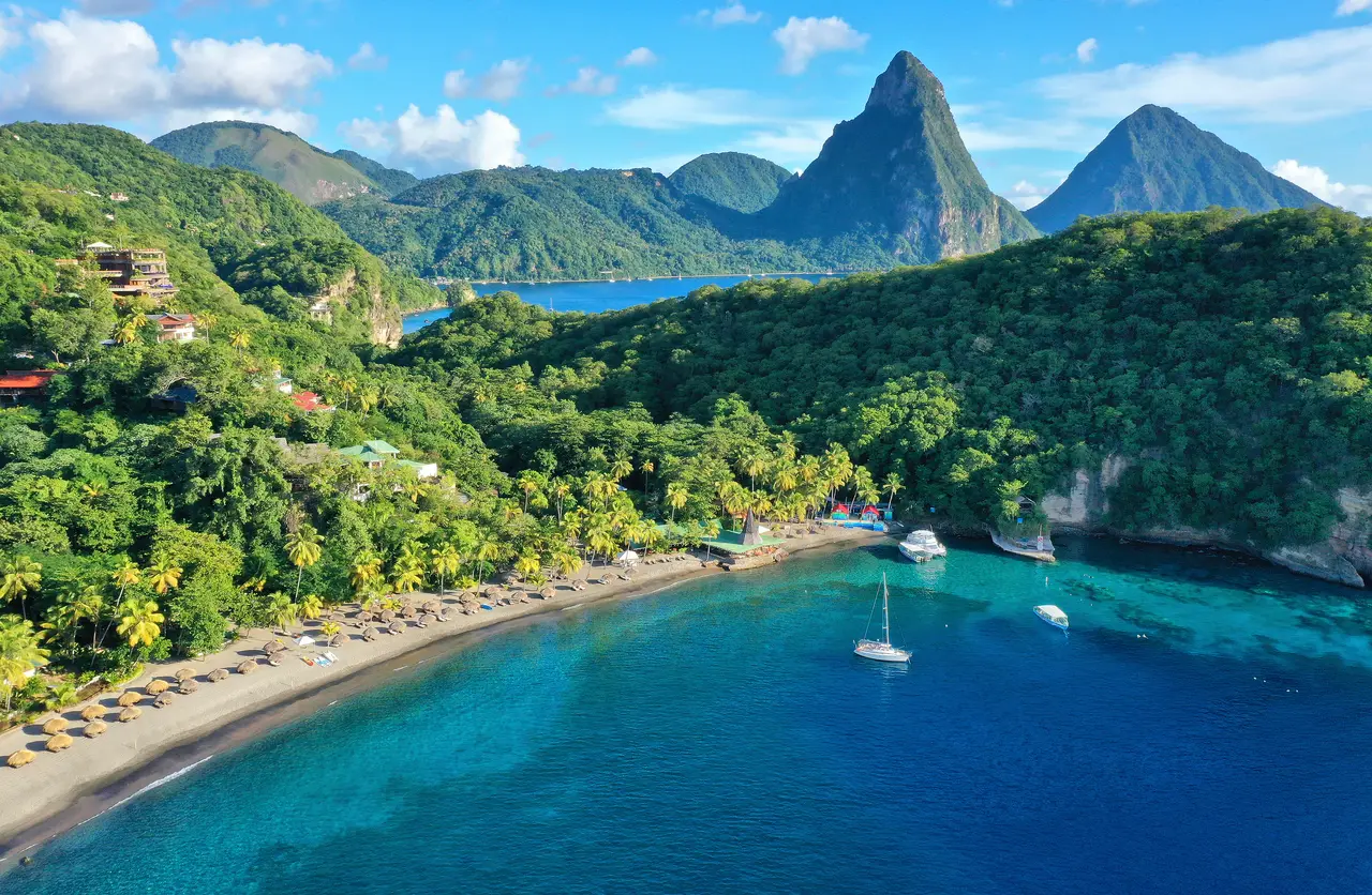 Anse Chastanet bay from the air: the dark-sand beach with its thatched parasols, boats at anchor, the forested hillside and the Pitons.