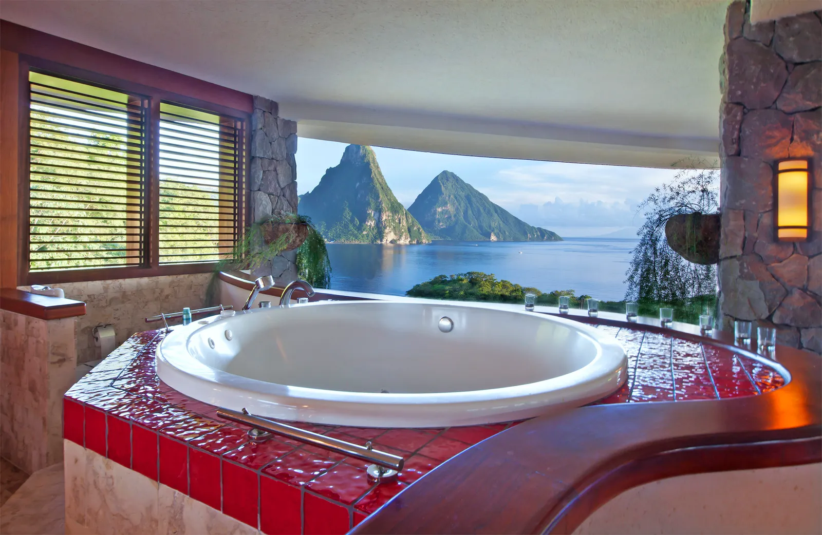 A sanctuary’s round whirlpool tub on a plinth of red tiles, timber louvres to the left and the Pitons in front.