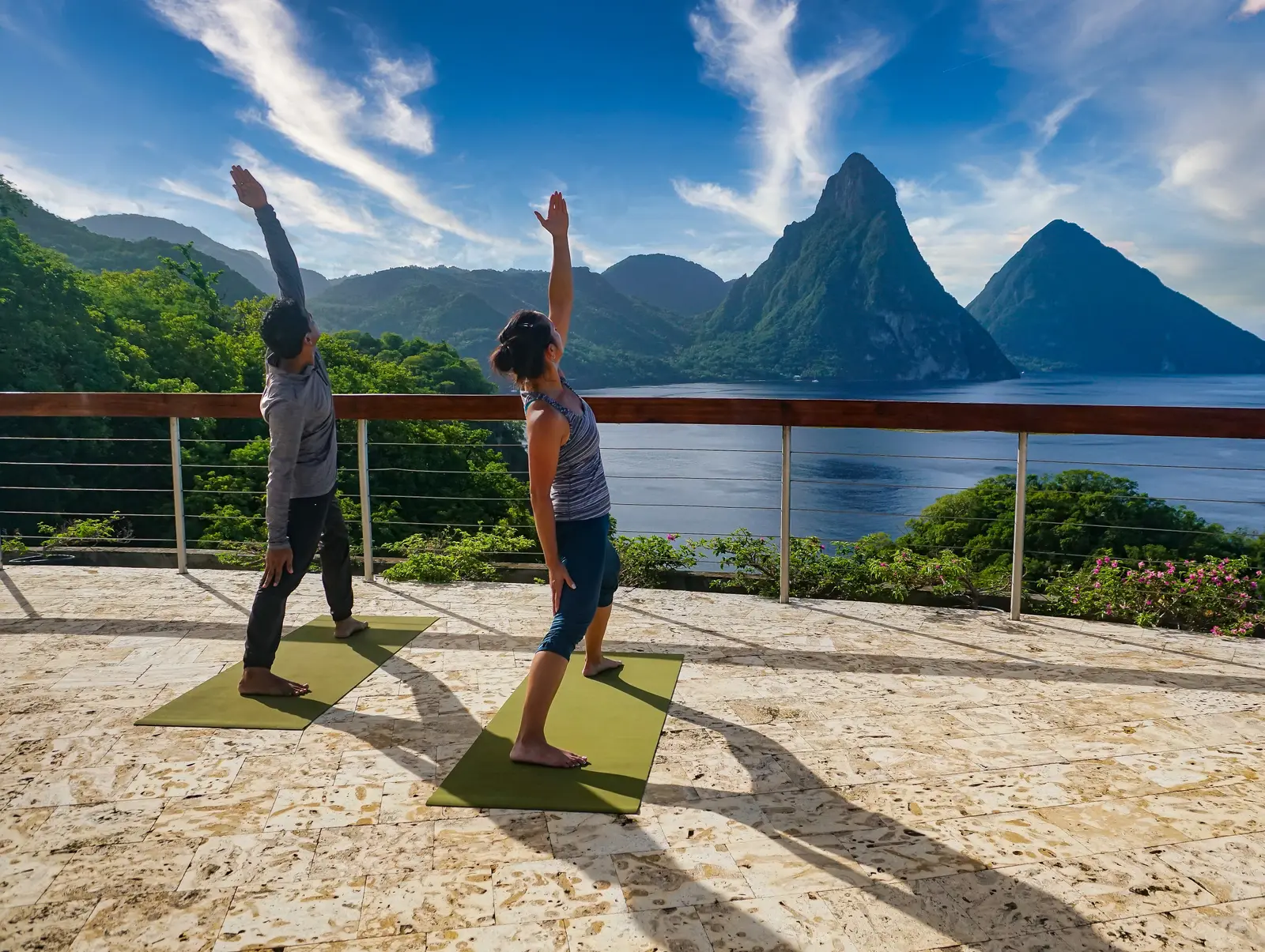 2 people doing yoga on mats on the Celestial Terrace, with the rail, the bay and the Pitons under a streaked sky.