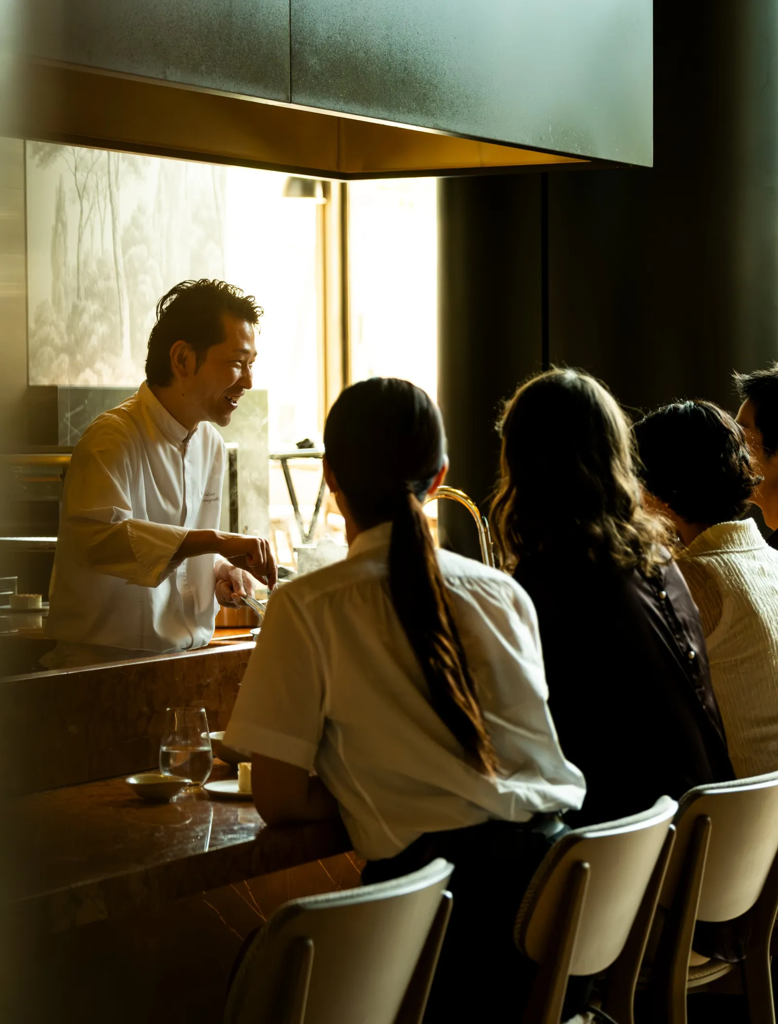 At Janu Mercato’s counter a cook talks with 4 seated guests, in the light from the window.