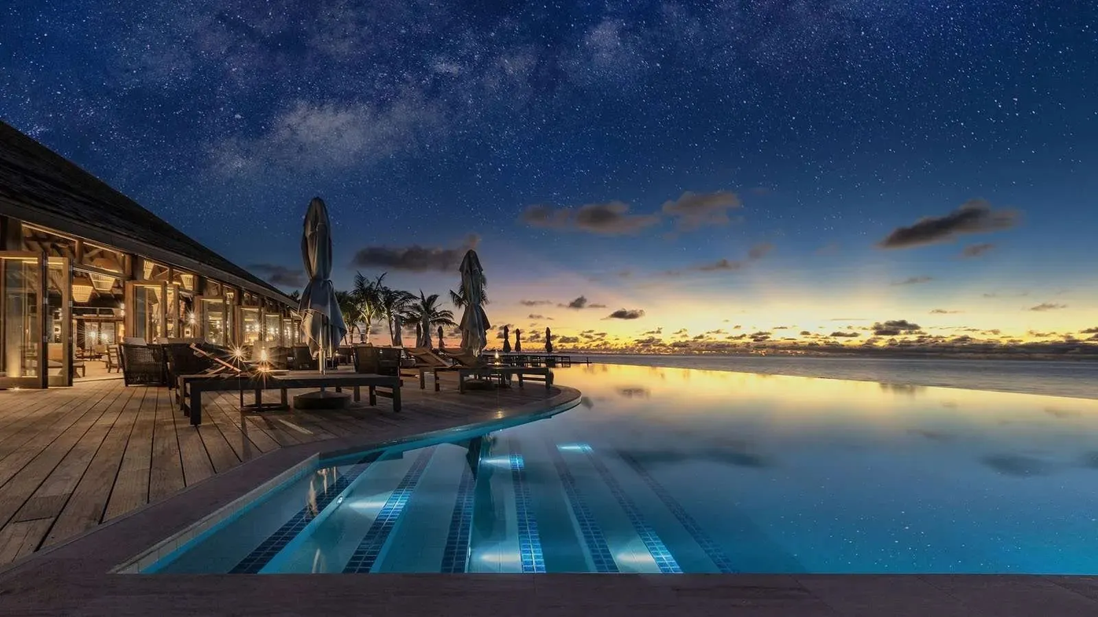 The infinity pool at night, lit up, with the restaurant lights and a starry sky