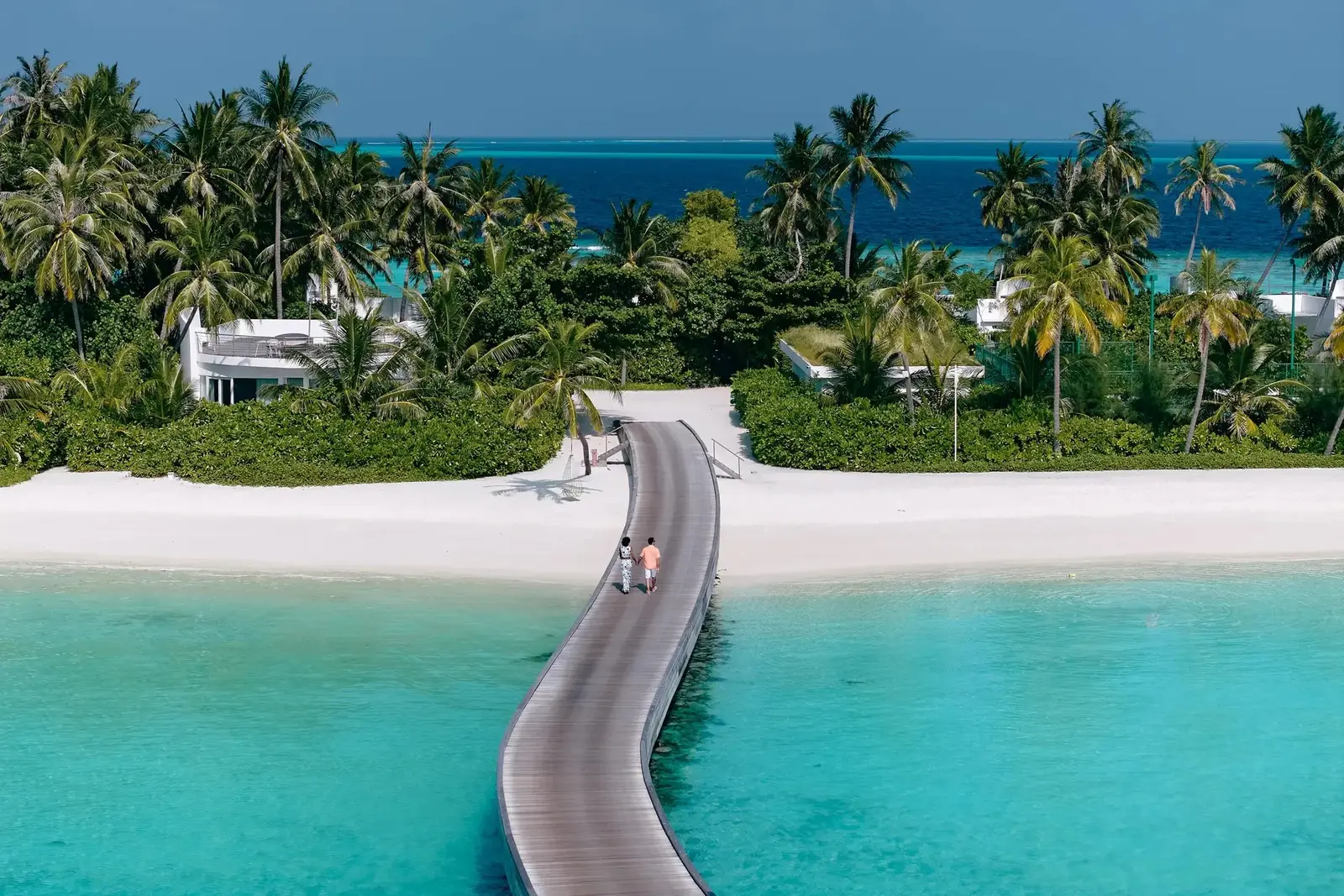 A curving timber walkway over the lagoon to the white beach and the palms, 2 people walking along it