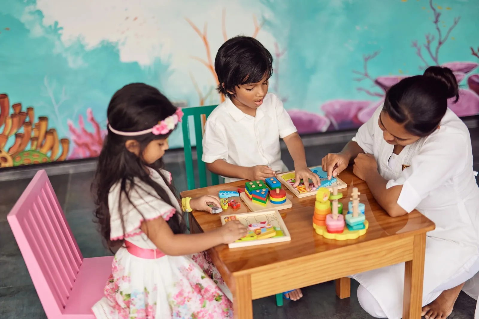 2 children and a carer at wooden puzzles, in front of a coral mural