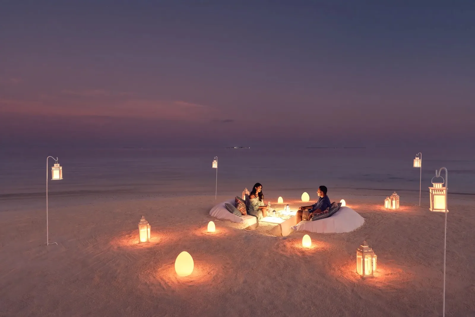 A couple dining on the sand at dusk, lanterns and small lights scattered around them
