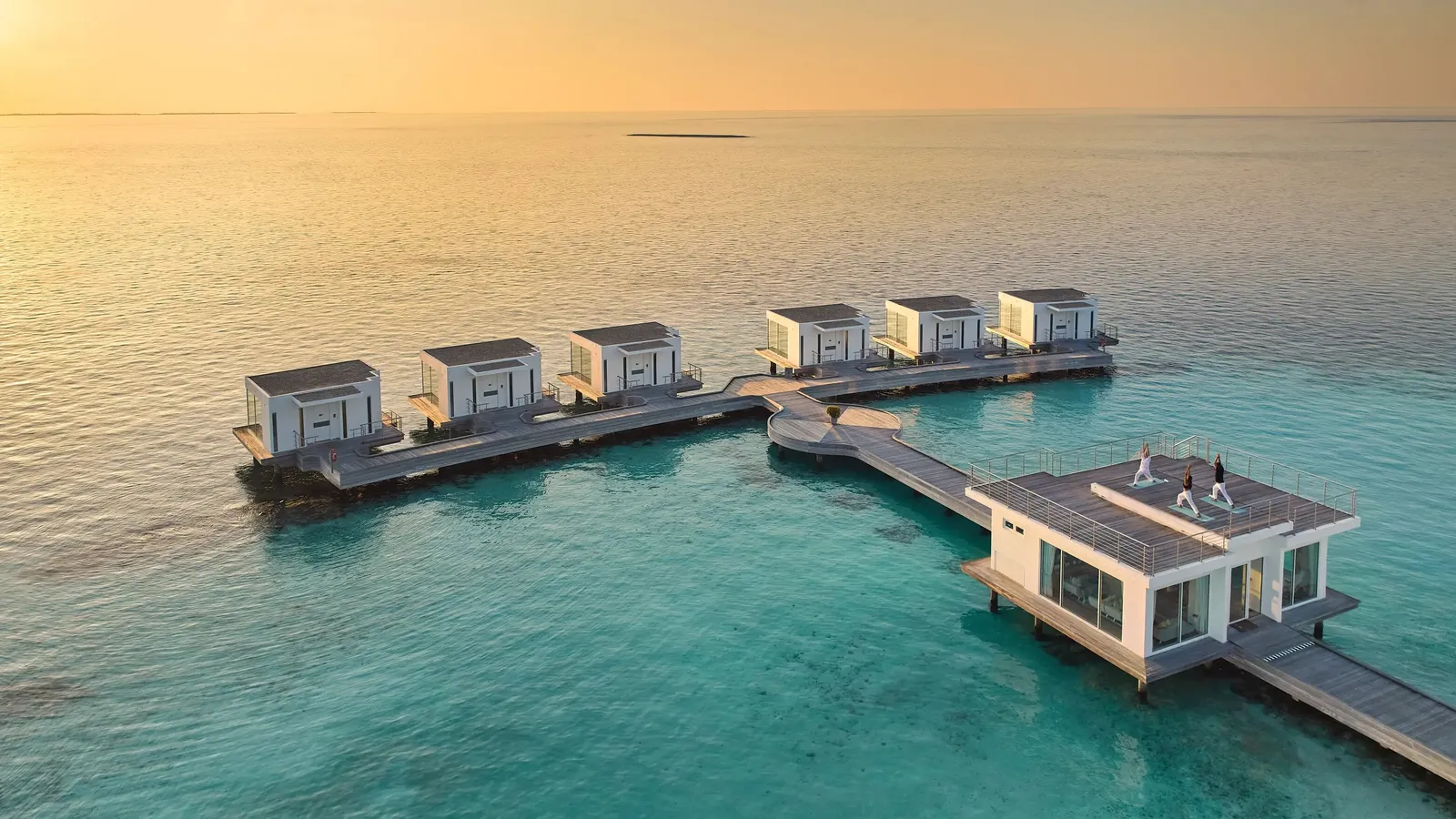 Talise Spa’s overwater pavilions at sunset, linked by a walkway, 3 people doing yoga on a rooftop