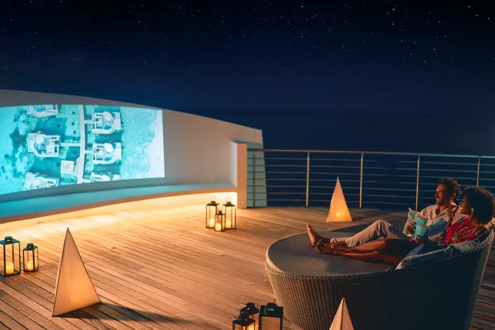 A couple on a round daybed on a rooftop terrace at night, among lit lanterns and an open-air cinema screen