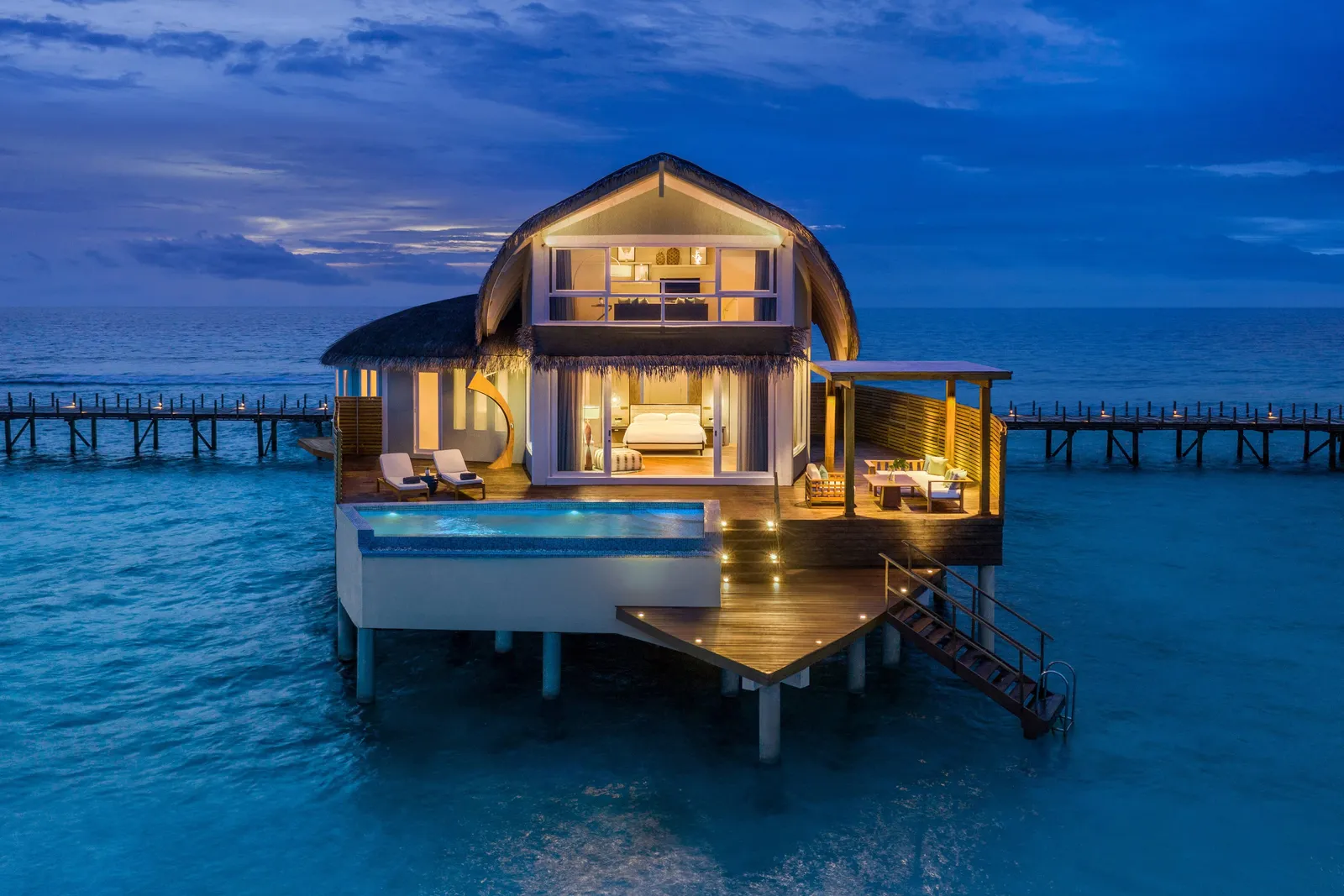 A Duplex Overwater Pool Villa at dusk, lit up, its deck pool and steps into the lagoon