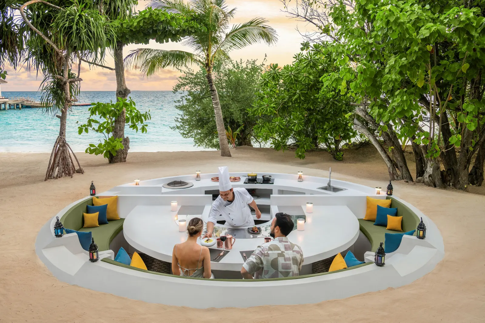 A chef serving a couple at RIHA’s table sunk into the sand, among the palms