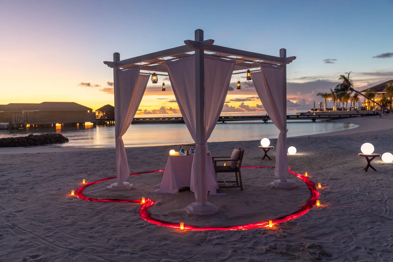 A table under a white gazebo on the beach at sunset, inside a heart of candles