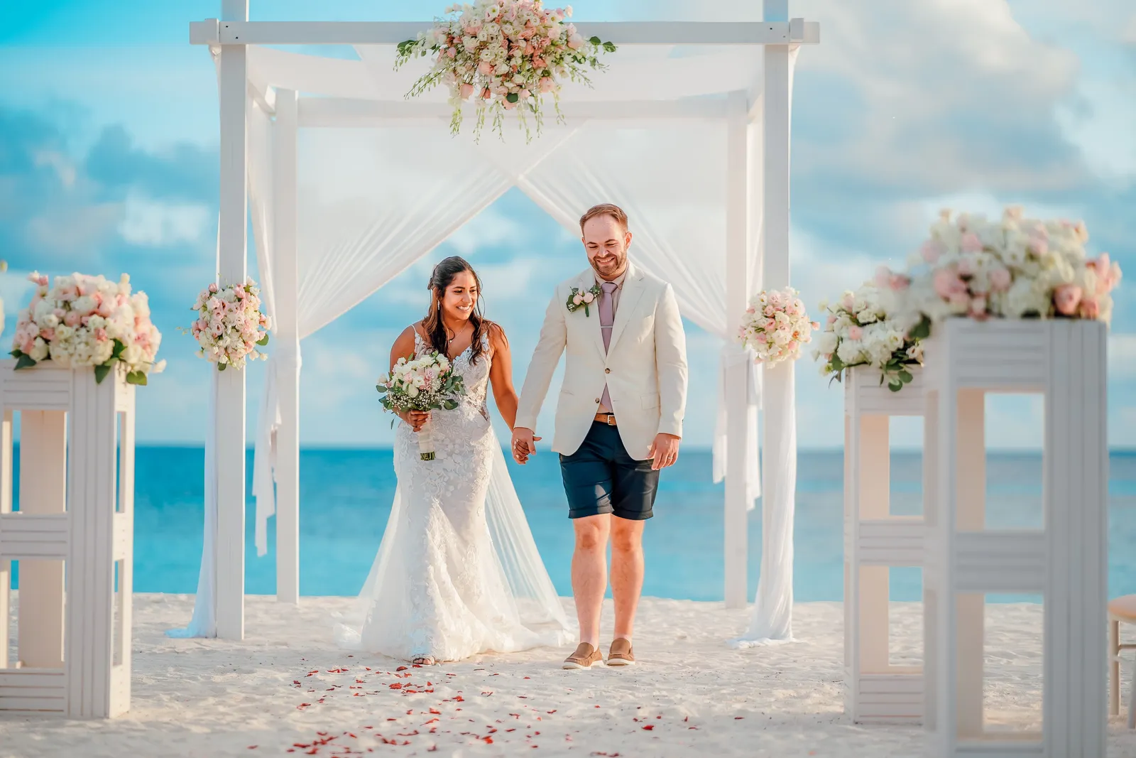 A married couple beneath an arch of white flowers on the beach