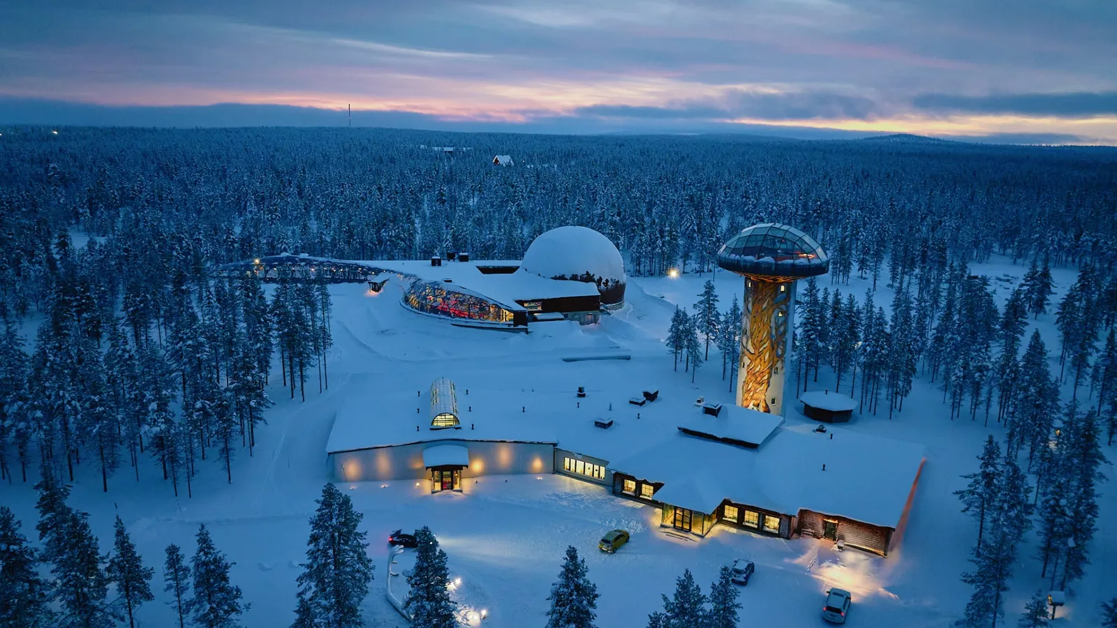The Kakslauttanen Cosmos complex from above at dusk: the planetarium dome, the Igloo Tower with its glass room on top, and the snowy forest.