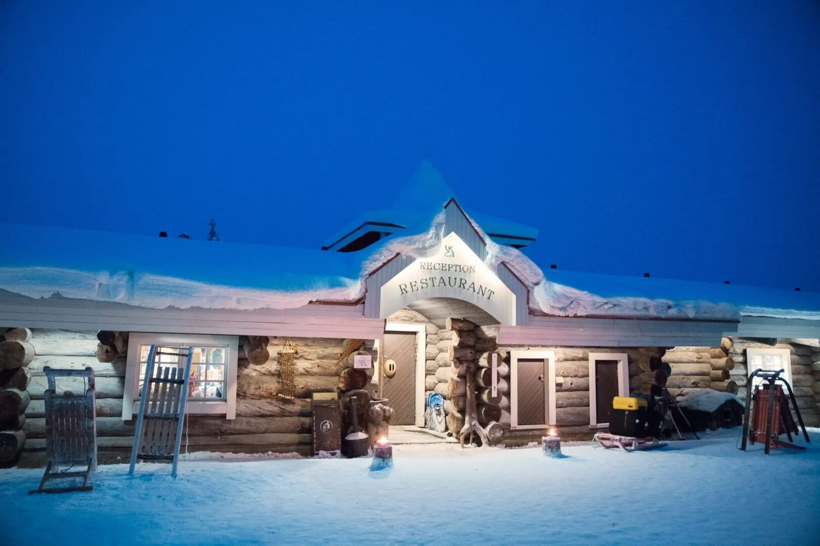 The log building of the East Village reception and restaurant at dusk, its sign lit and kick-sledges leaning against the wall.