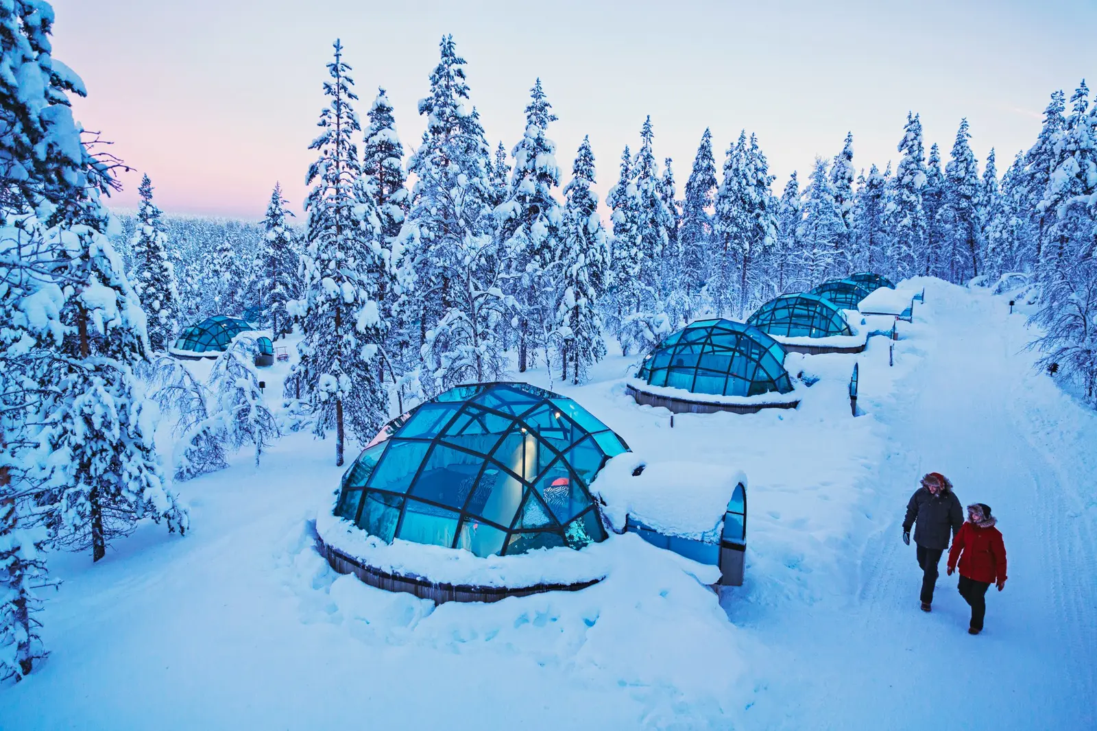 2 people walk through the snow beside a row of blue-green glass igloos, among snow-laden spruces under a pink sky.