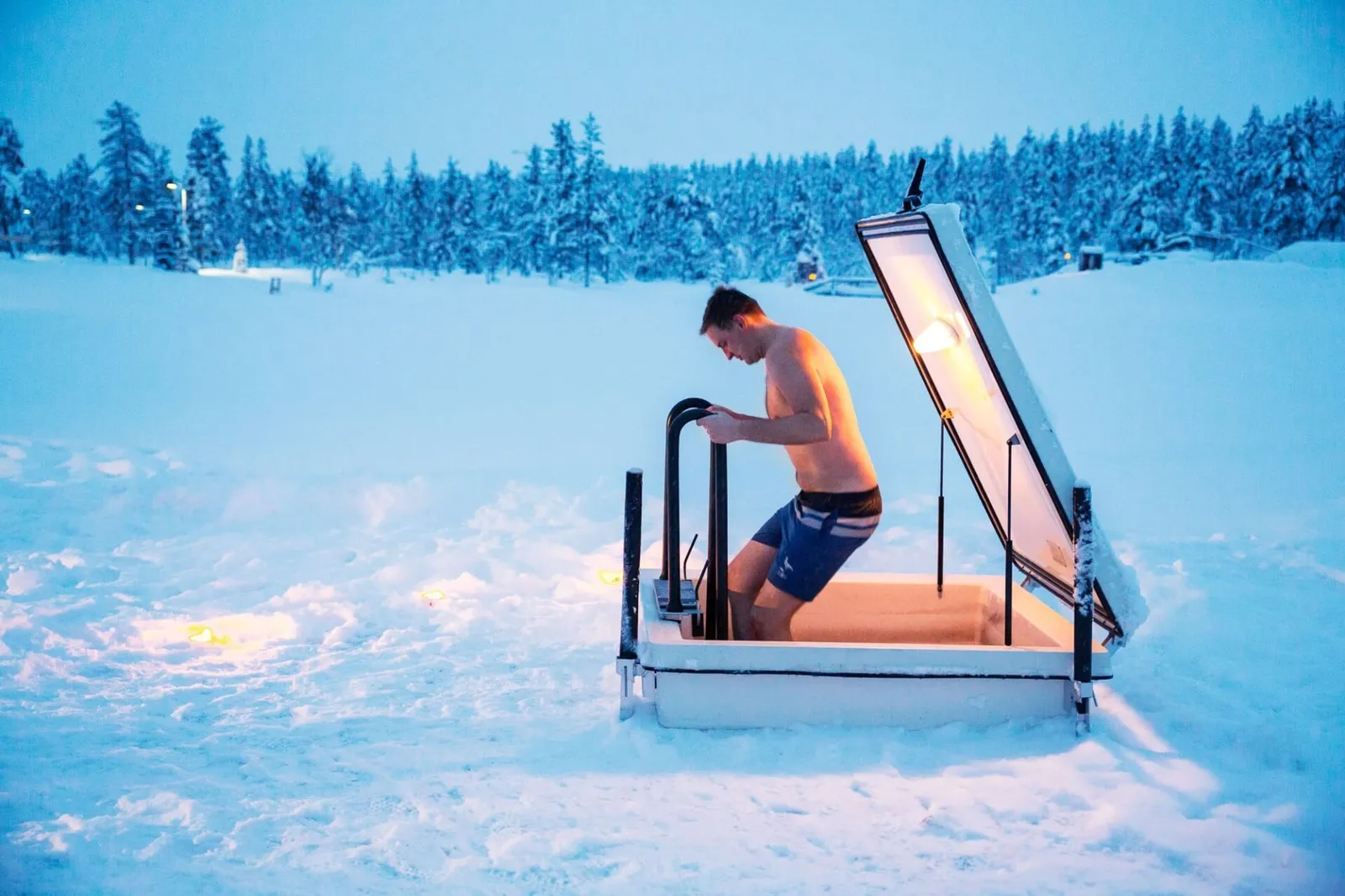 A man in swimming shorts climbs out of the ice pool, its lid raised, in the middle of the snow; the forest behind.
