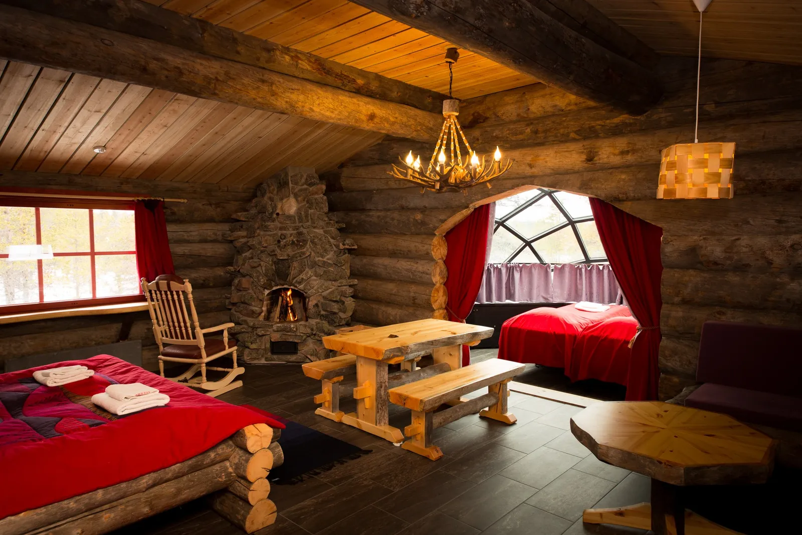 A Kelo-Glass Igloo: the log room with its stone fireplace and wooden table and, beyond the arch with red curtains, the bed under the glass dome.