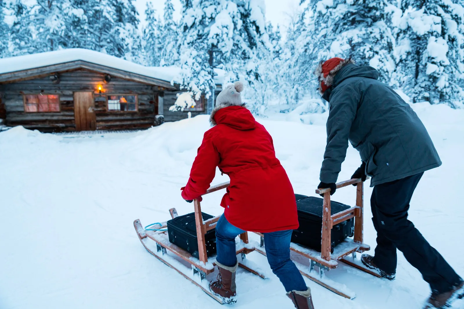 2 guests push wooden kick-sledges loaded with suitcases across the snow towards a log cabin.
