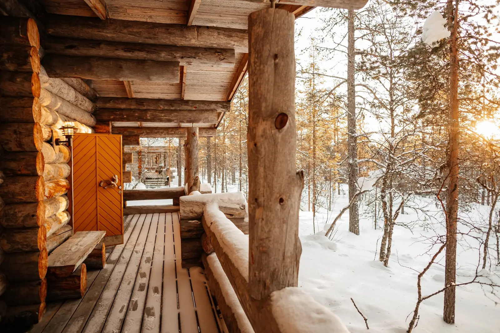 The porch of a log cabin with an orange door and a bench, and the low sun between the pines in the snow.