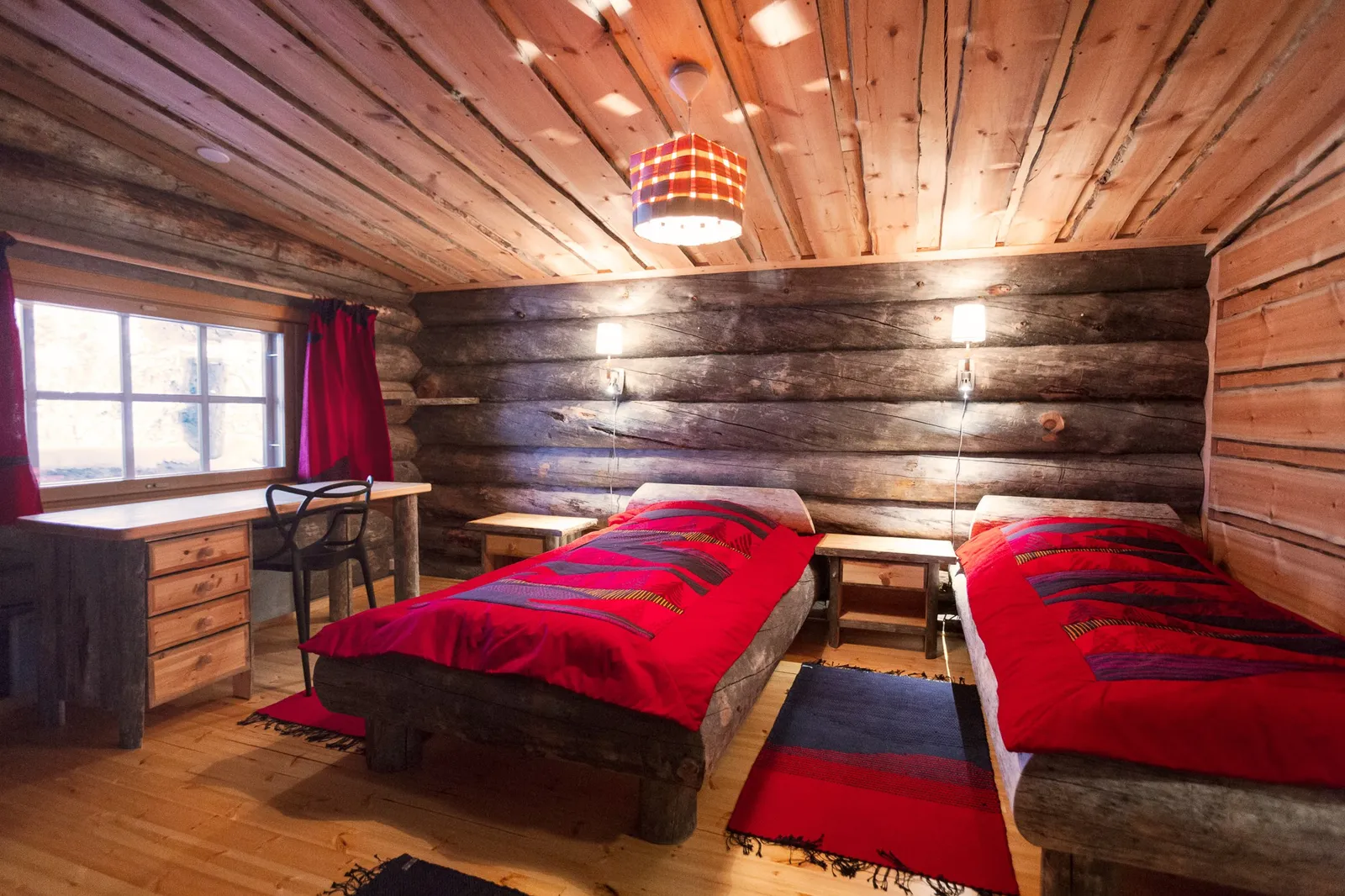 A bedroom in a Large Cabin: 2 wooden beds with red covers, a desk under the window and log walls.