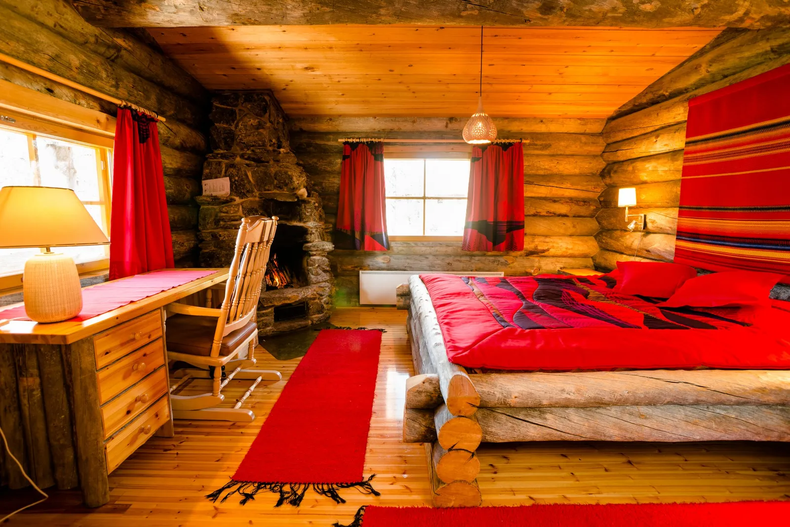 Inside a Small Cabin: the double bed with a red cover, the stone fireplace in the corner and a desk with a lamp.