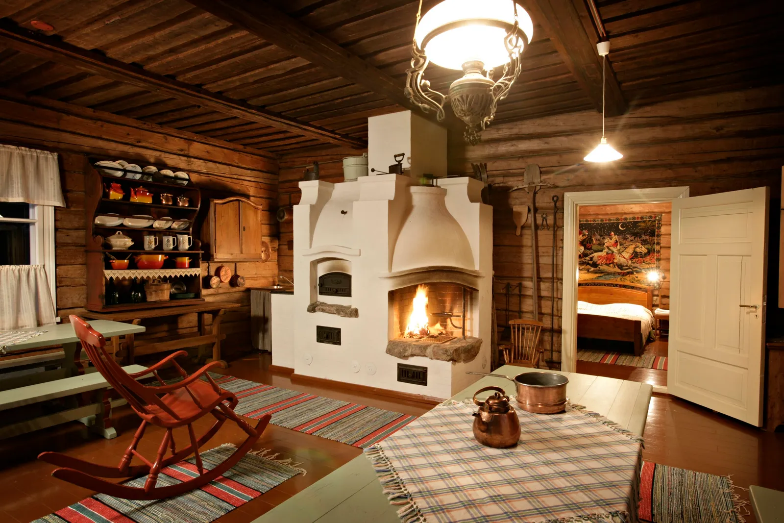 The Traditional House: the white masonry stove with a fire burning, the rocking chair and a dresser of crockery.