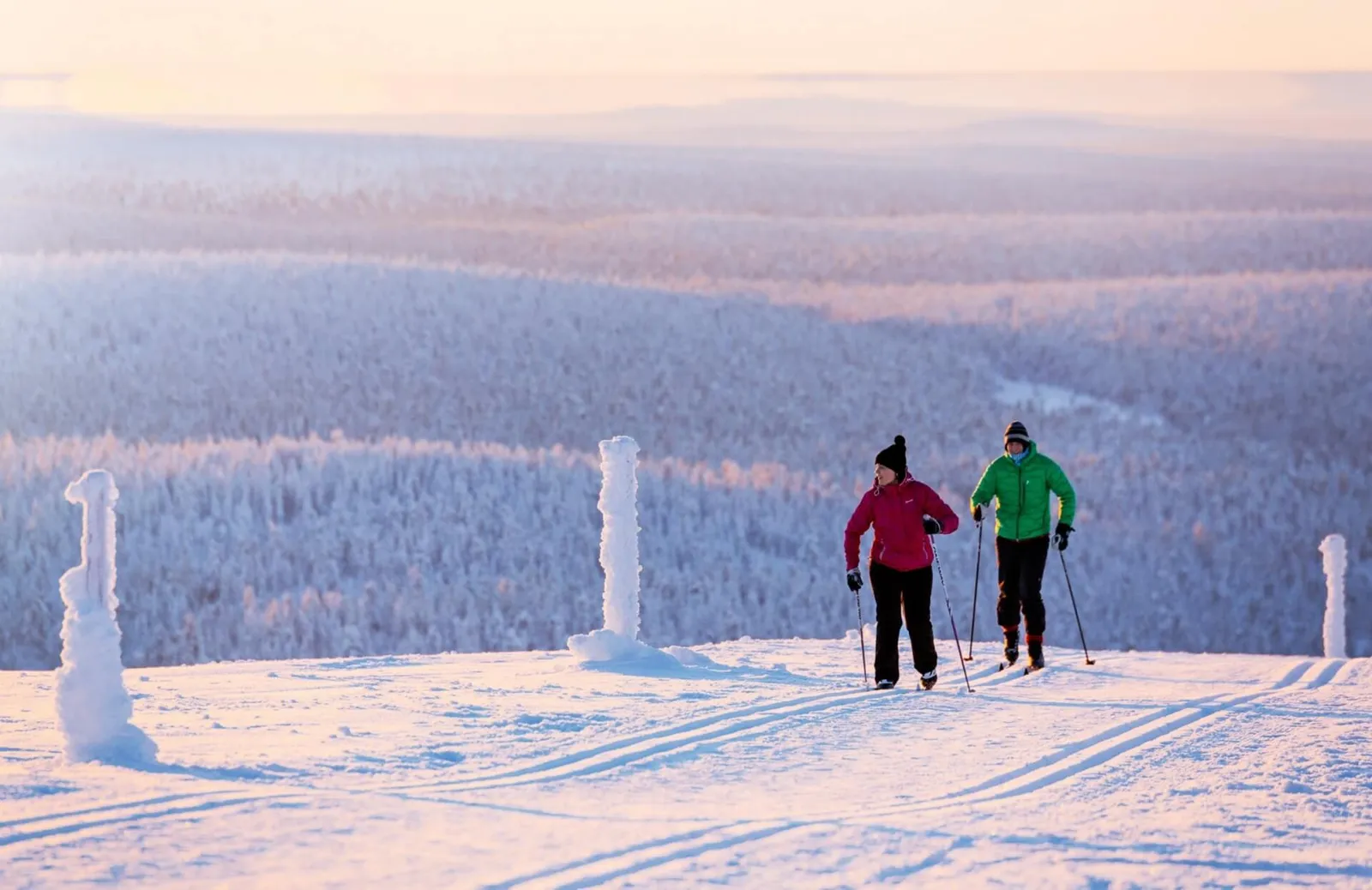2 cross-country skiers on the groomed track on a hilltop, the snowy forest running to the horizon in pink light.