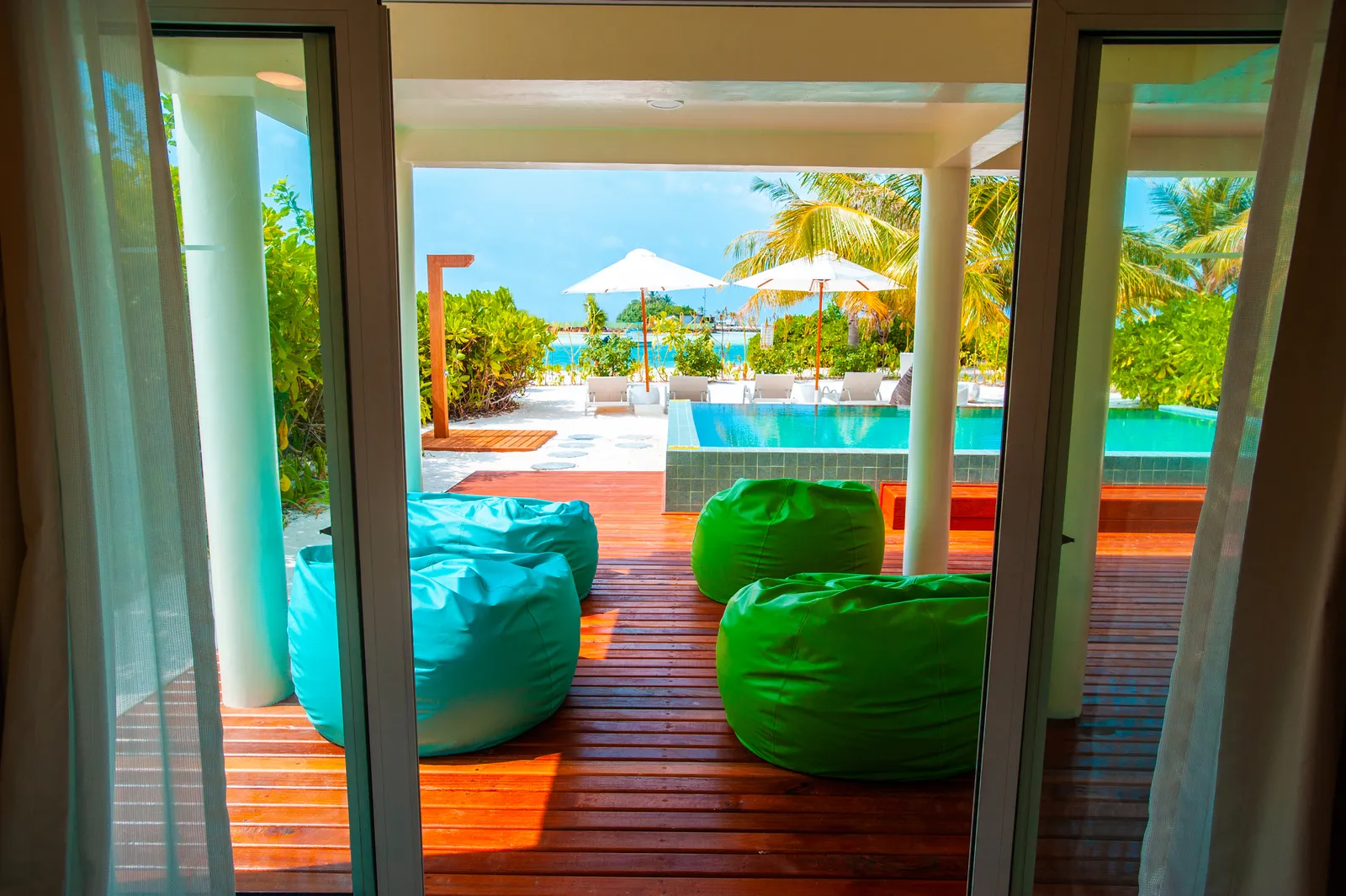 From the living room of a 3-bedroom villa: bright poufs on the deck and the pool beyond the door.