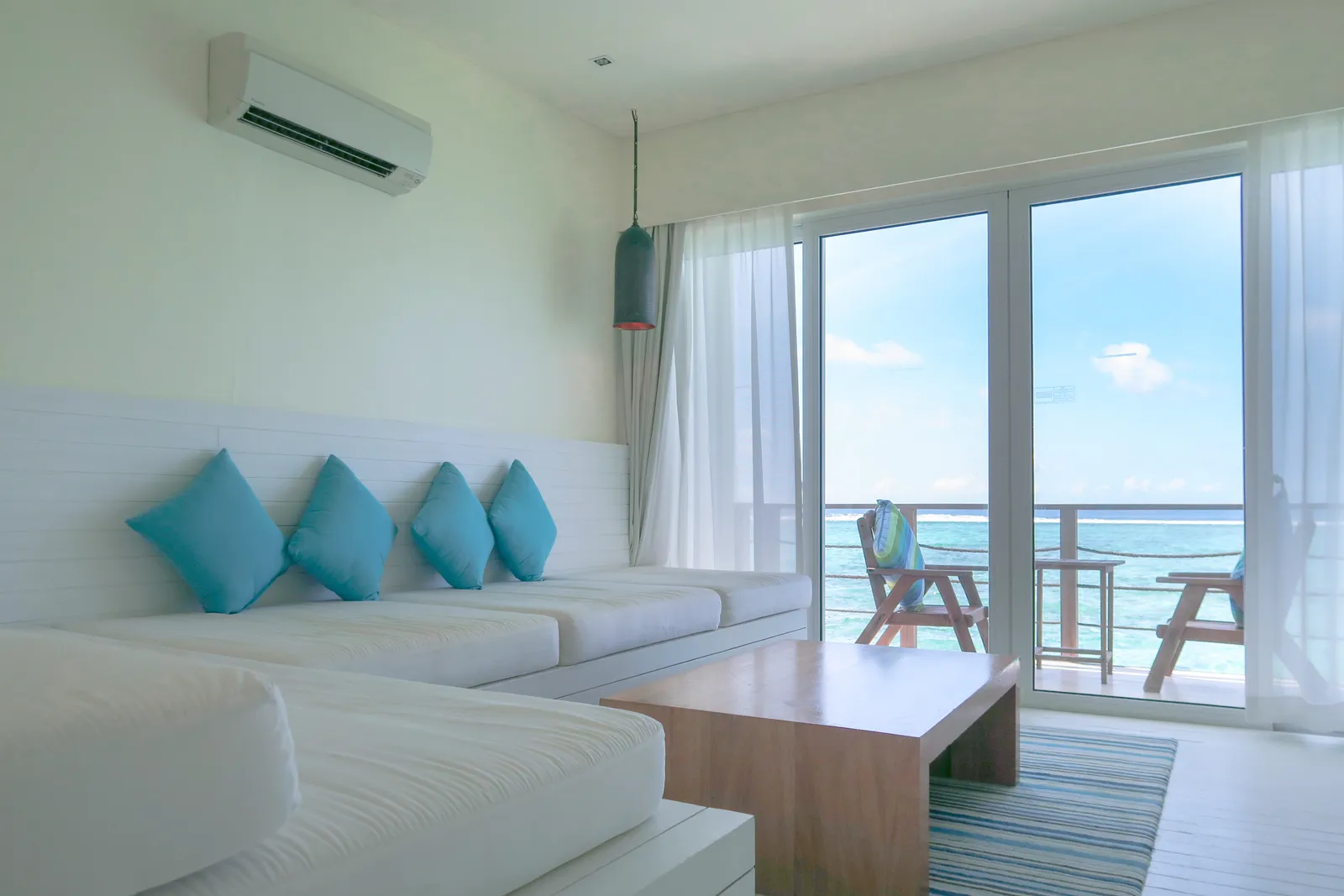 A water villa’s living room: the white sofa with turquoise cushions and the glass doors onto the balcony and the sea.