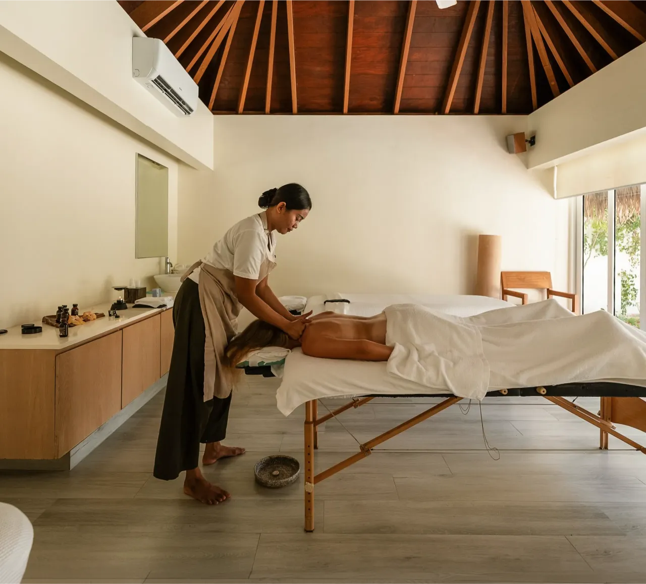 A spa room with a timber ceiling: a therapist begins a massage for a guest on the table.