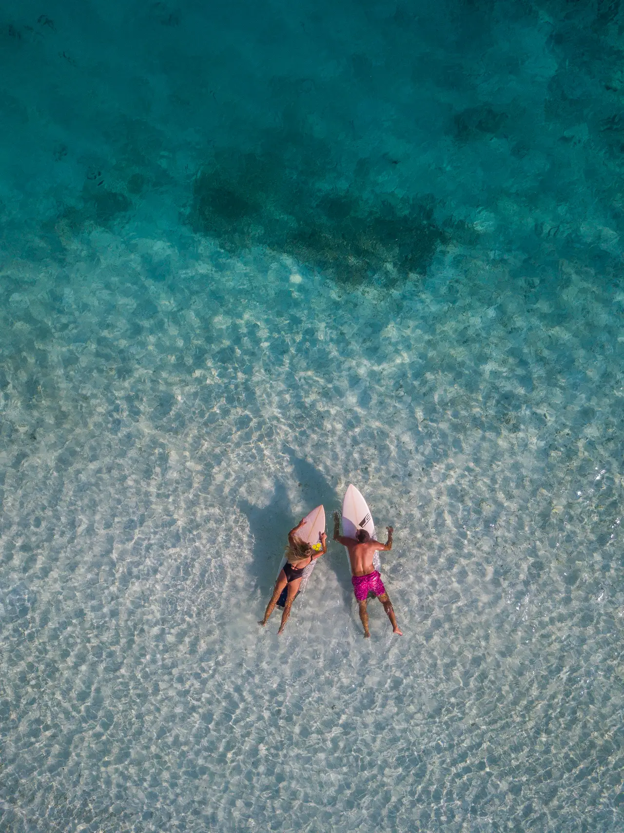 2 surfers lying on their boards in shallow, clear water, seen from above.