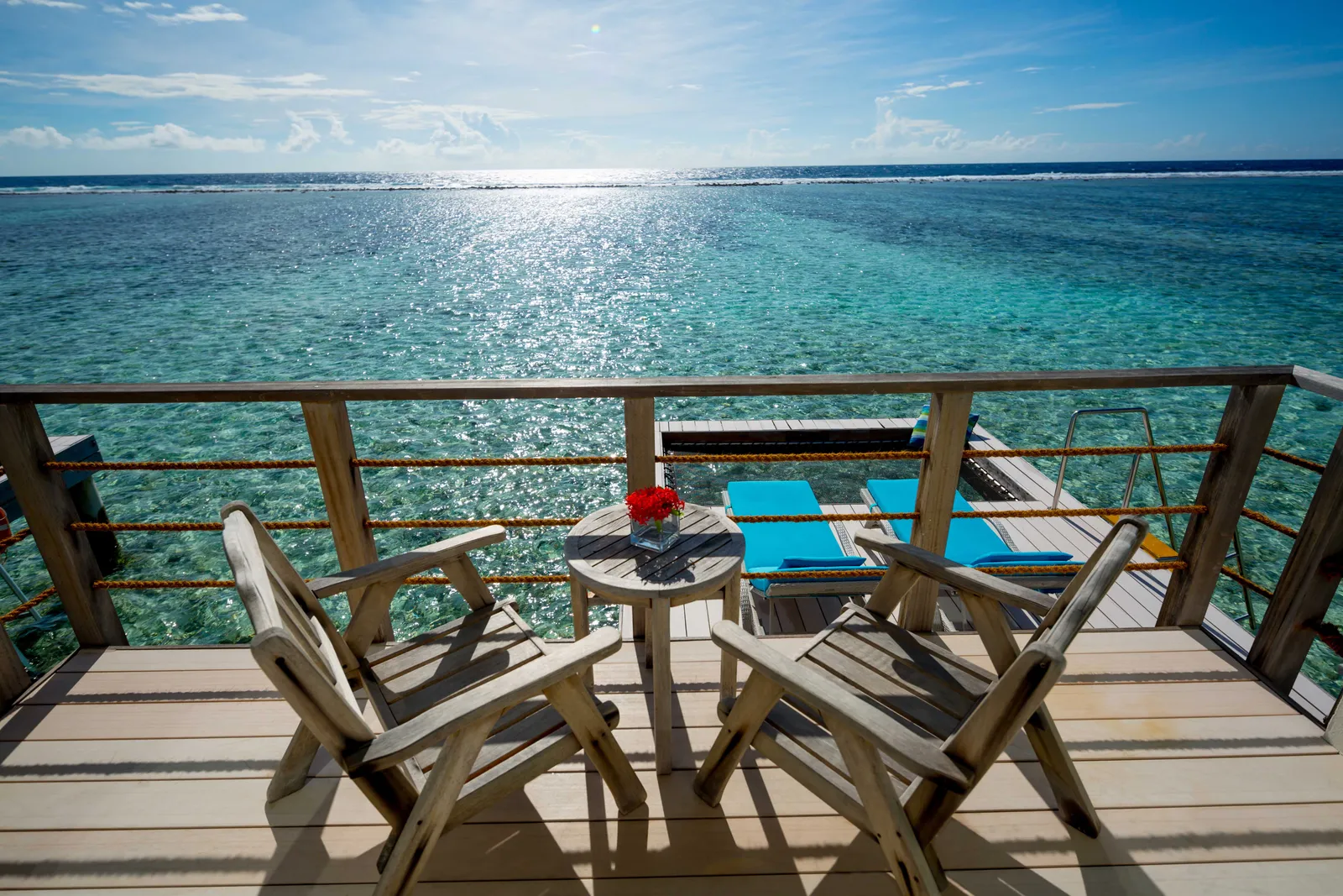 The deck of a water villa: 2 wooden chairs, the loungers and the open sea glittering.