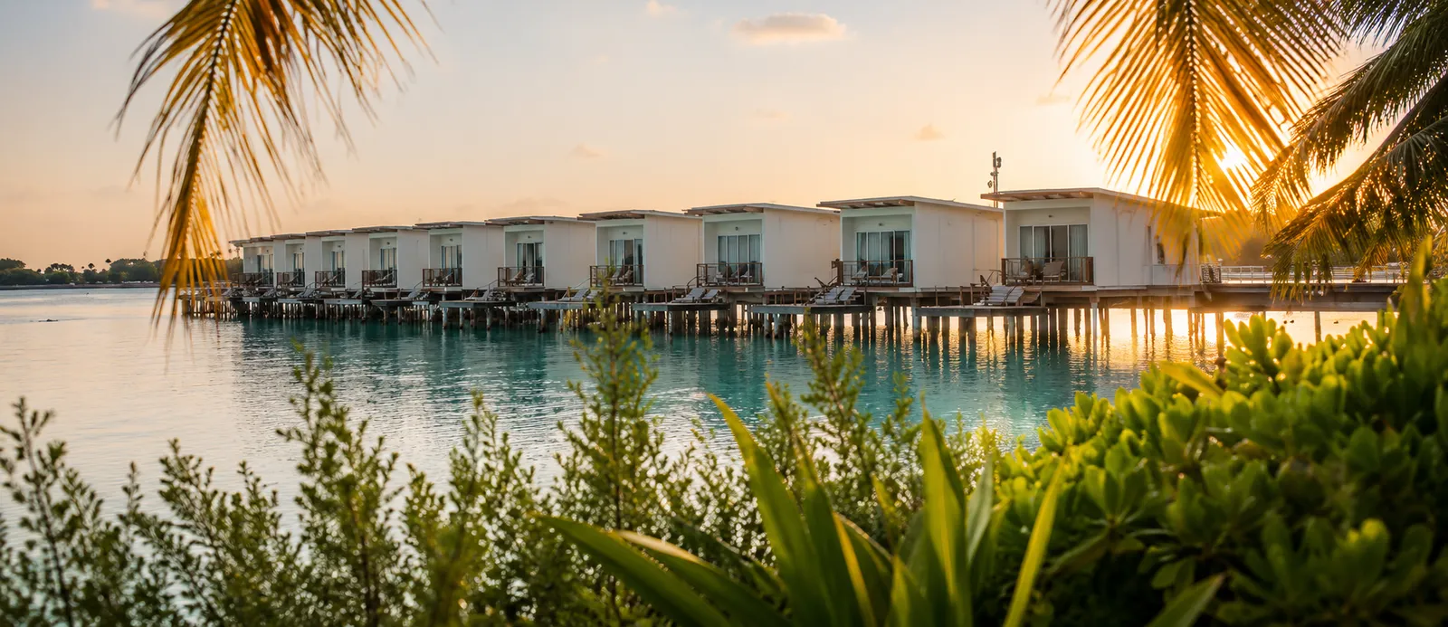 The water villas at sunset, seen from the shore through palm fronds.