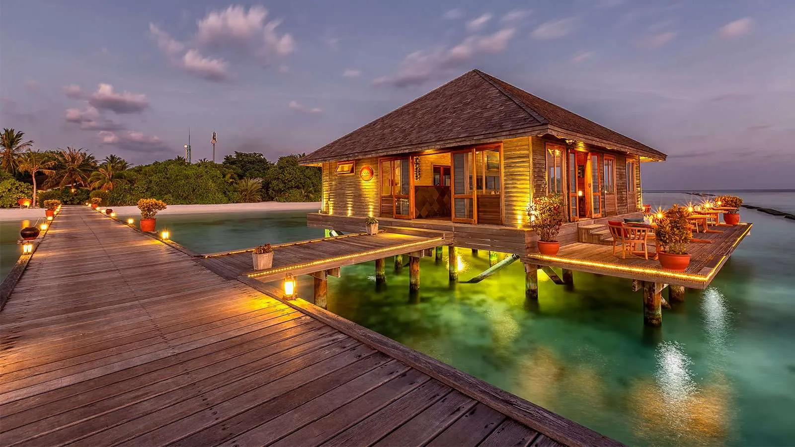 Aqua at dusk: the restaurant on stilts lit up at the end of its jetty, with the lagoon glowing green beneath.