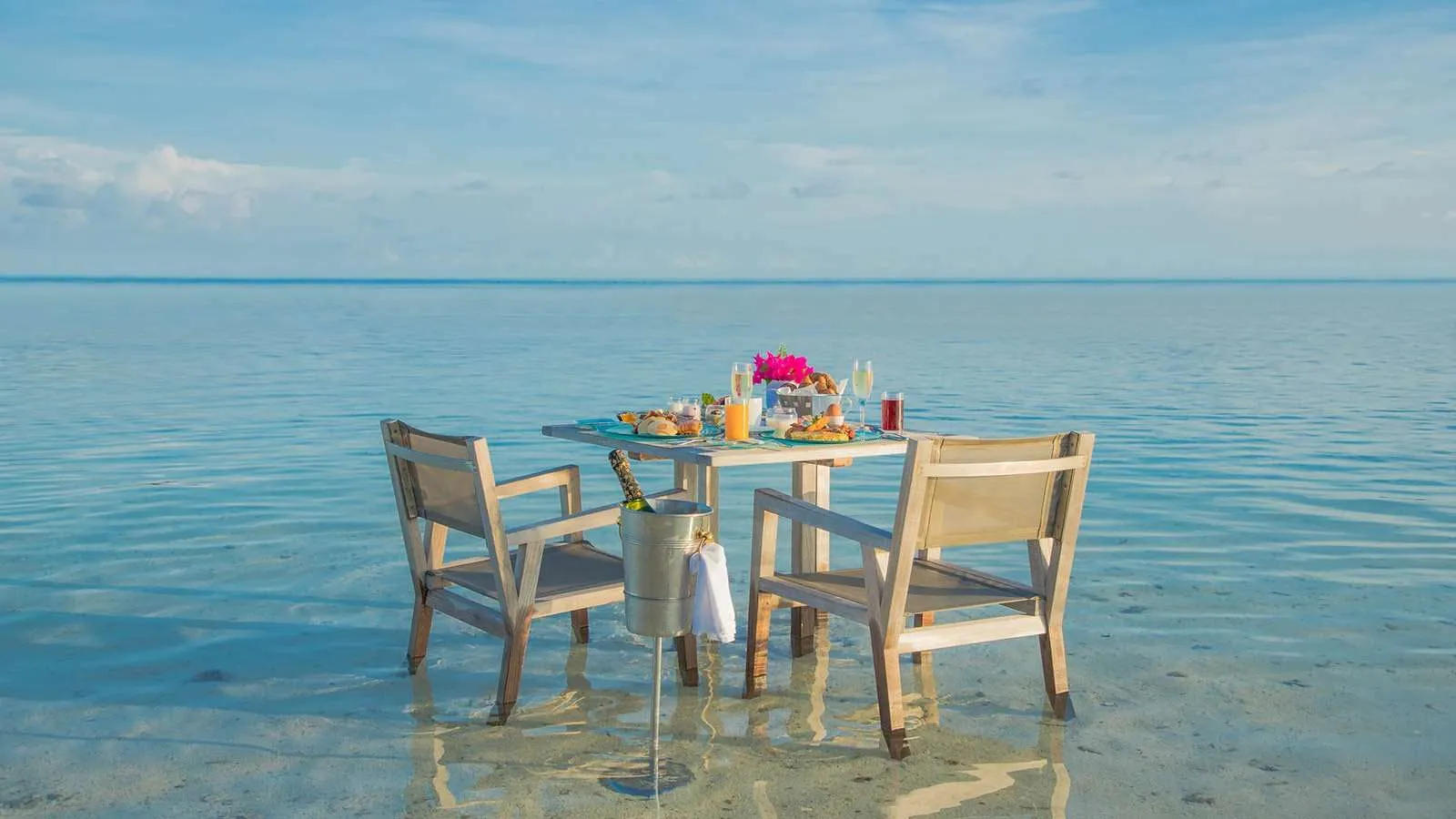 A breakfast table laid in the shallow water of the lagoon, with 2 wooden chairs.