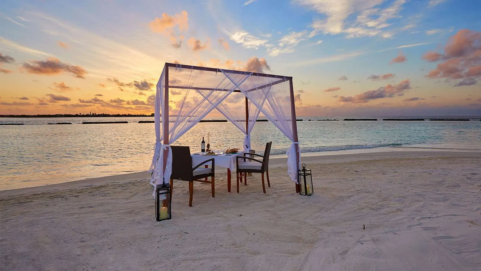 A table for 2 under a white canopy on the beach at sunset, with lanterns lit.