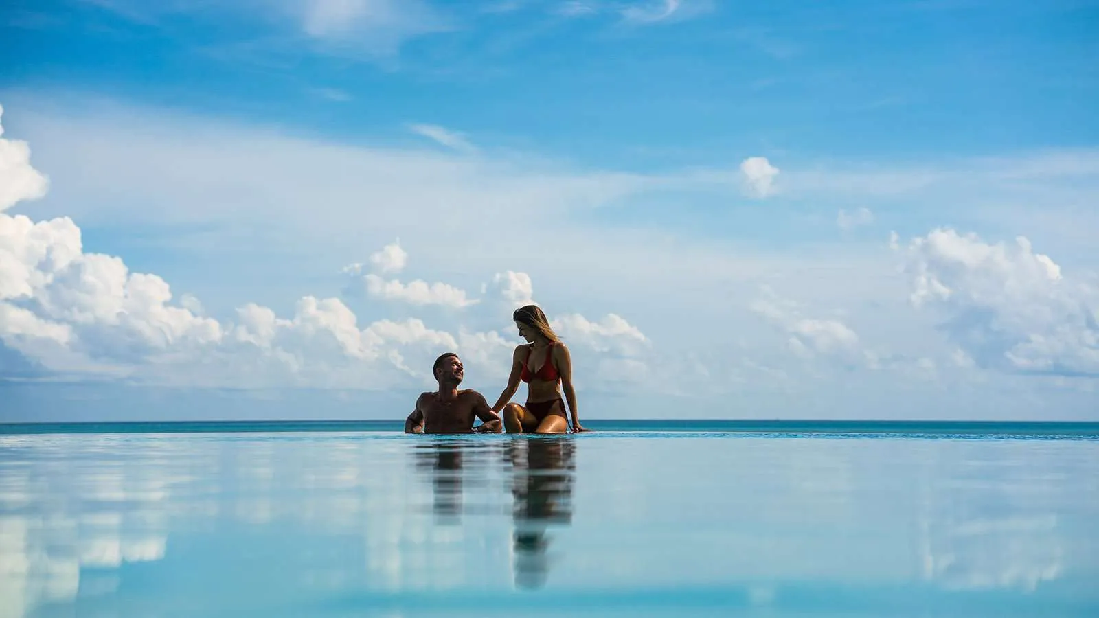 A couple leaning on the edge of the infinity pool, the sea flat and the clouds reflected in the water.