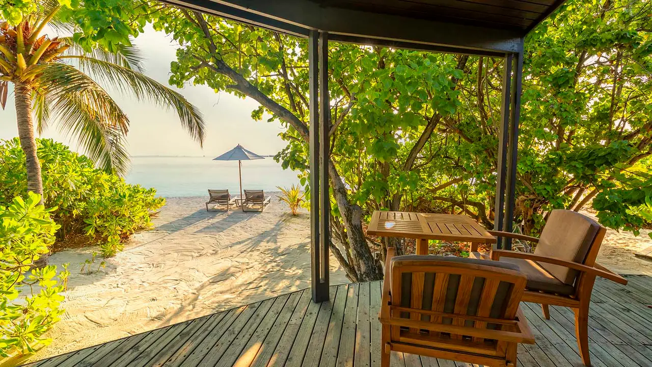 A Jacuzzi Beach Villa’s wooden veranda, with its table and chairs, the trees and the beach with loungers ahead.