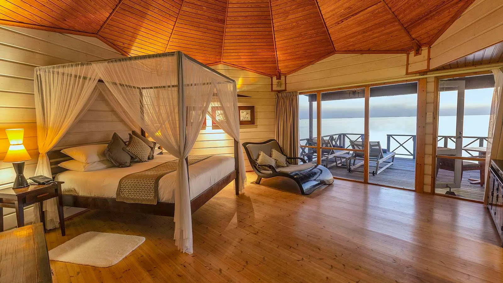 A Jacuzzi Water Villa bedroom: the four-poster bed, the wooden floor and the glass doors onto the deck and the sea.