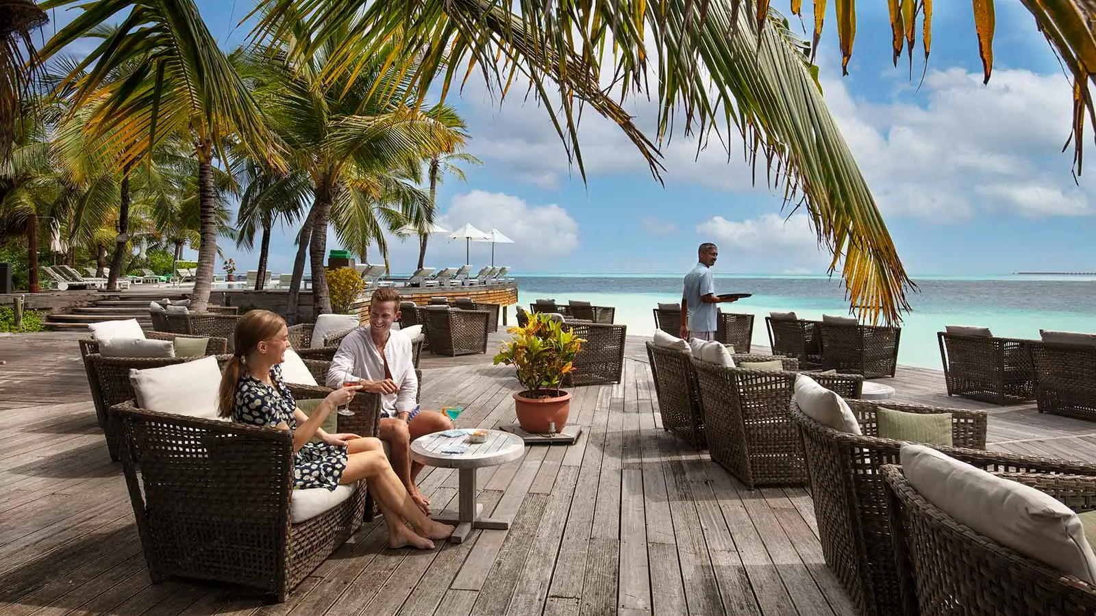A couple in wicker armchairs on the Kandu Bar deck, in the shade of the palms, with the lagoon behind.