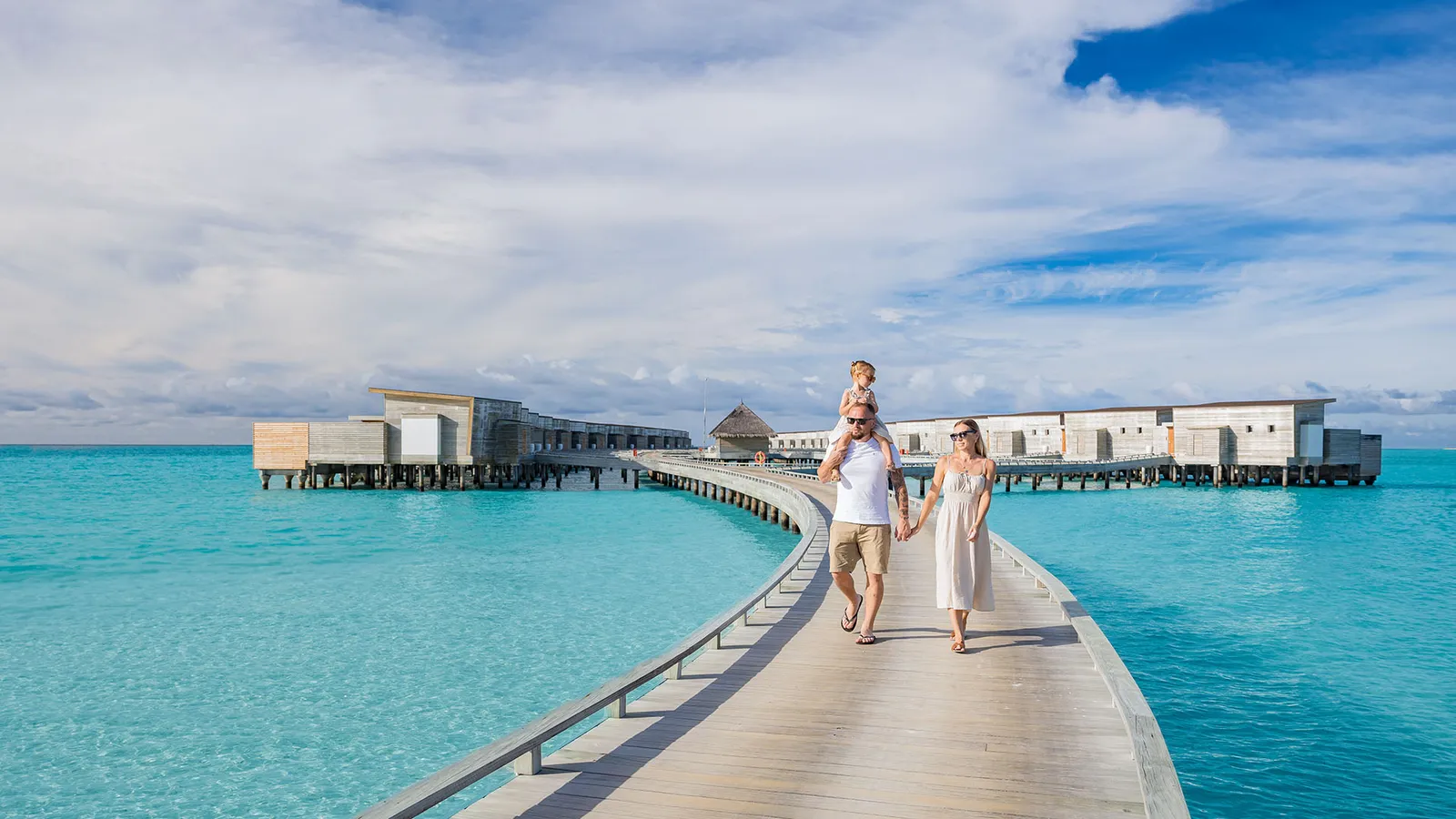 A family walking along the water-villa jetty, the child on his father's shoulders.