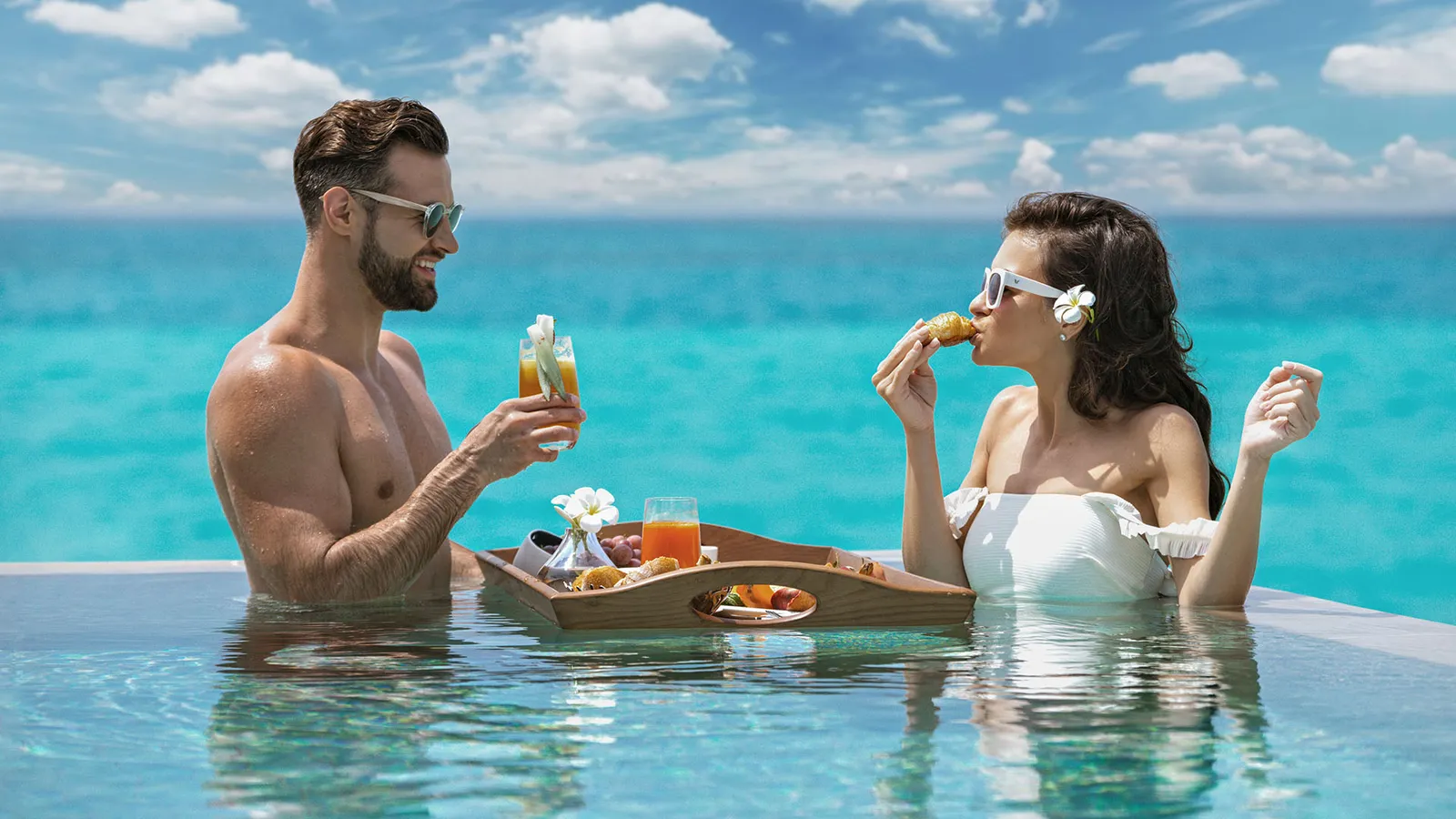A couple having breakfast in their private pool, the tray floating and the sea behind them.