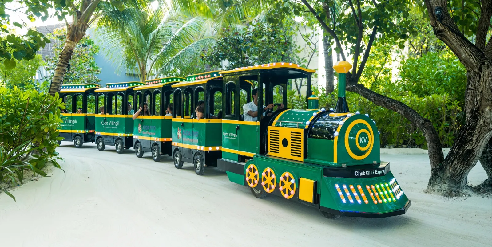 Kuda Fiyo's green and yellow island train on the sandy path under the trees.