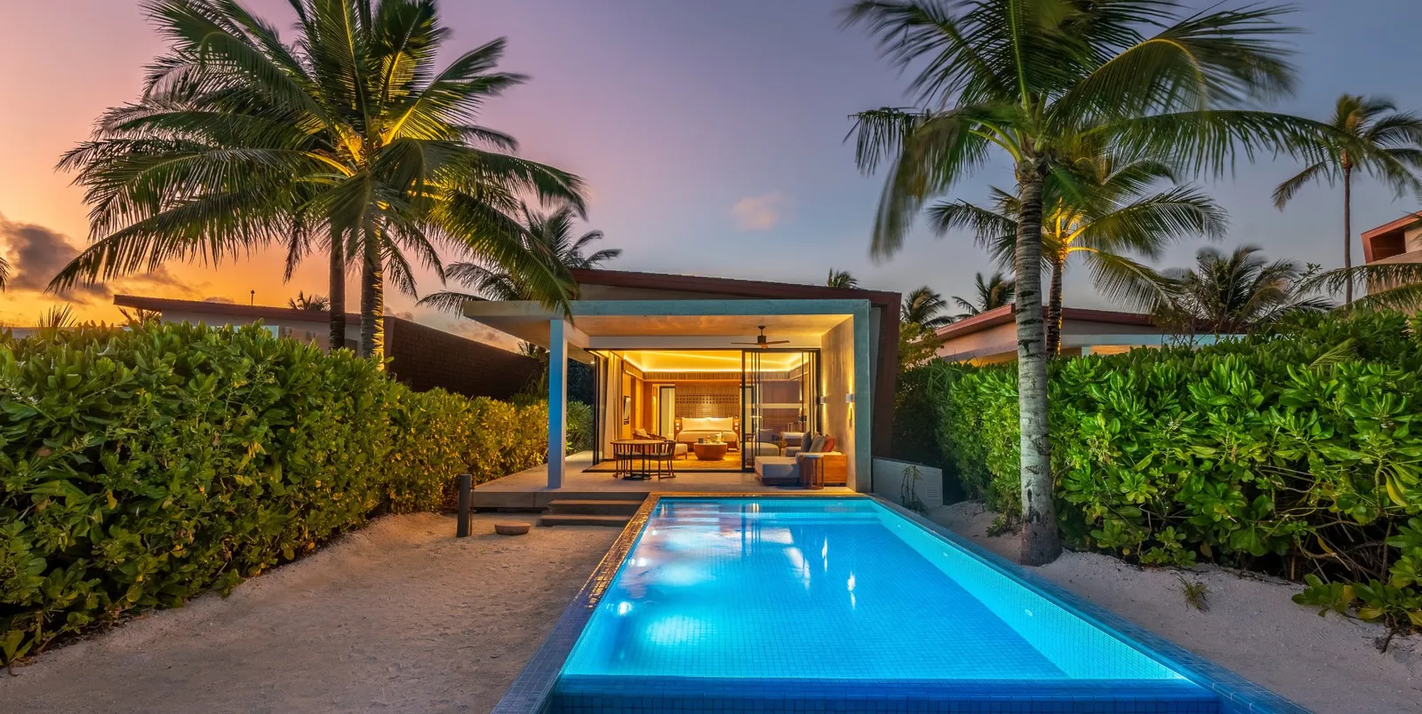 A Sunset Beach Villa at dusk, the lit bedroom at the end of the pool among the palms.