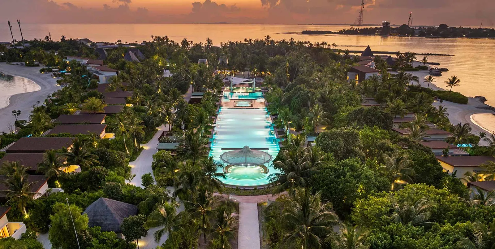 The central pool at dusk, lit up, with a table laid at the water's edge and the villas among the trees around it.