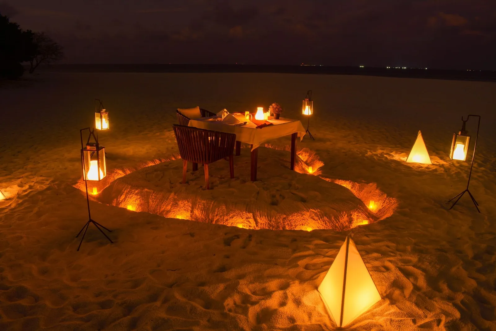 A private dinner on the sand at night: a table for 2 inside a ring of lanterns.