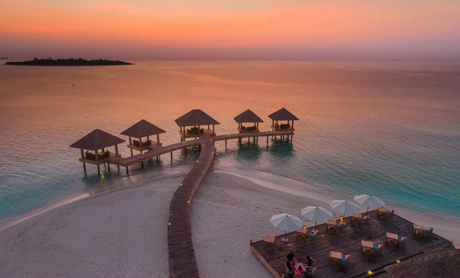 A walkway running from the beach to the gazebos on the water at sunset, with a parasol-shaded deck below.
