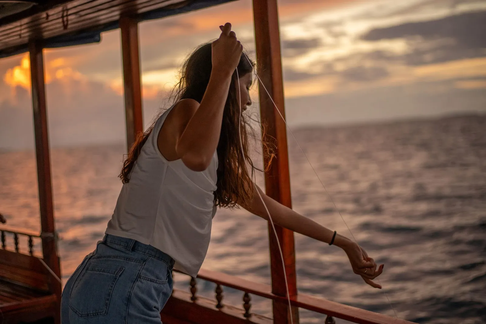 A young woman hauls in a hand line from a dhoni’s rail at sunset, over a grey and pink sea.